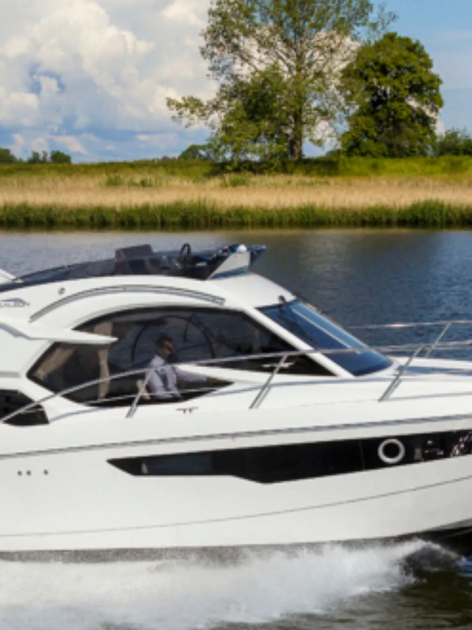 Een witte motorboot met zwarte ramen vaart over water, met een man aan het roer, langs een groene oever. — Galeon 300 Fly