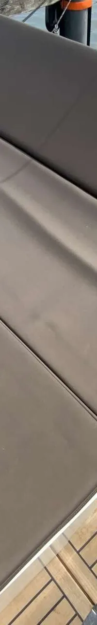 De foto toont een bootdek met bruine kussens en een houten vloer, met water en een andere boot op de achtergrond.