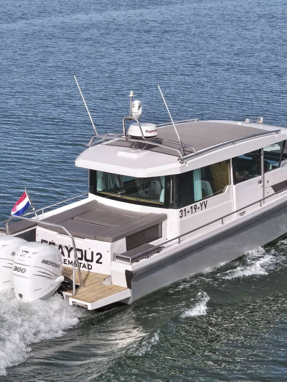 Een grijze motorboot met witte opbouw vaart op een waterweg, met een groene oever en blauwe lucht op de achtergrond.