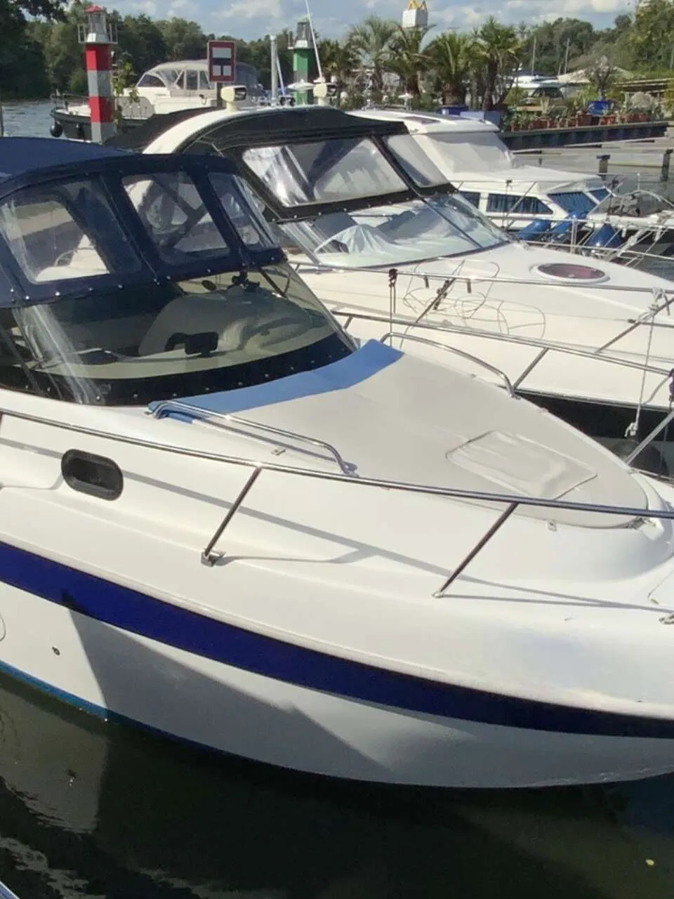 Een witte motorboot met blauwe strepen en een donkerblauwe kap ligt in het water, met andere boten op de achtergrond.