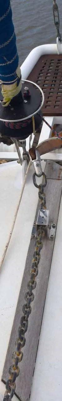 De boeg van een witte zeilboot met een blauwe rolfokhoes, ankerketting en reling, liggend aan een rotsachtige kust.