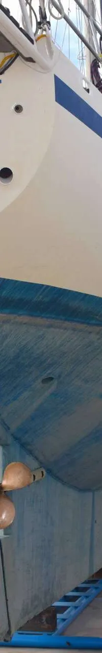 Een witte zeilboot met blauwe antifouling en de naam "ALEA IACTA EST HAMBURG 173488 S" op de spiegel, staat op bokken naast…