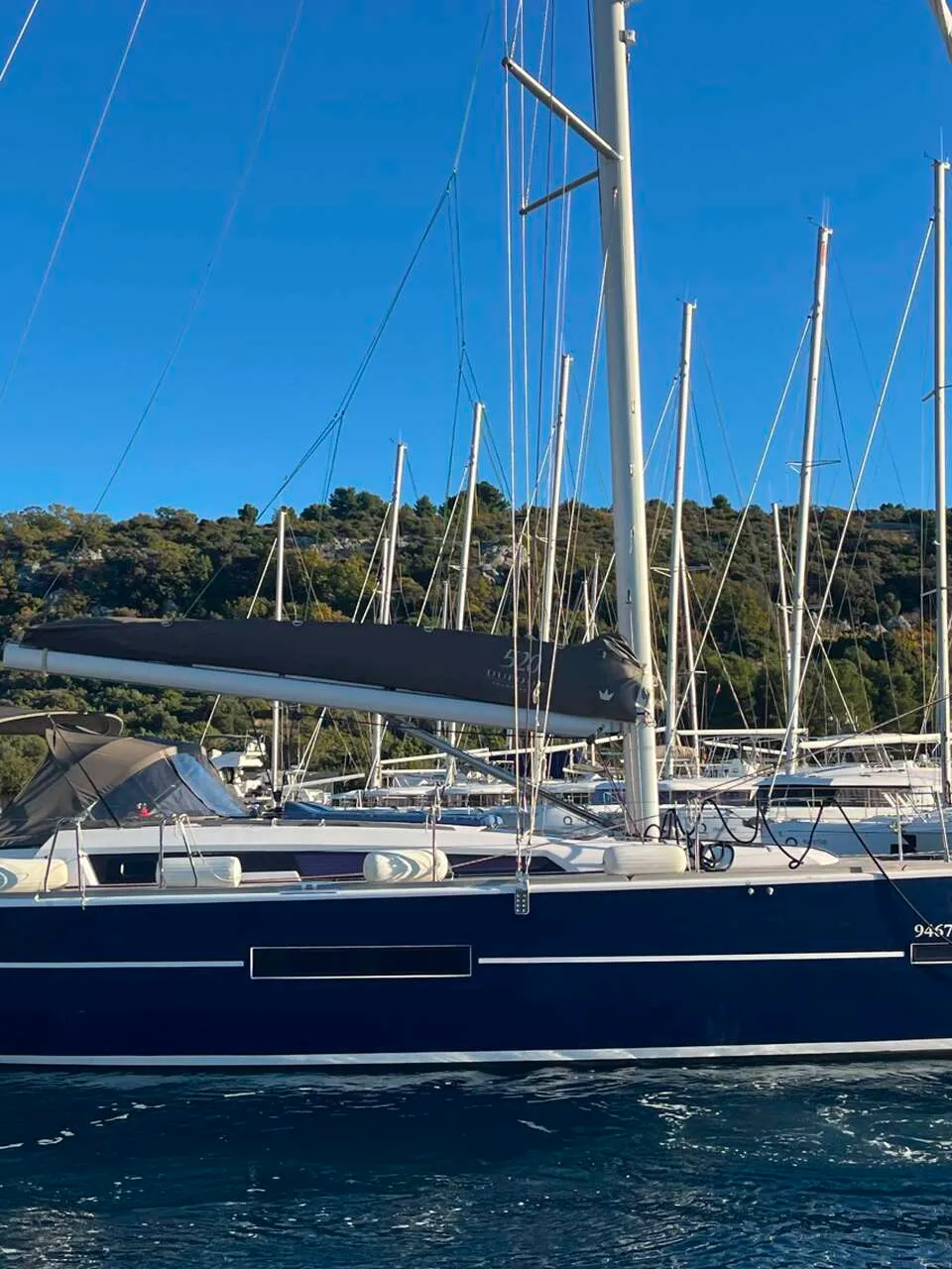 Een donkerblauwe zeilboot met een rode en witte streep op de spiegel ligt in het water, met een groene heuvel op de…
