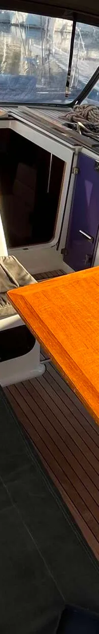 De foto toont een bootkuip met een houten tafel, banken met kussens, een kajuitingang en een buiskap.