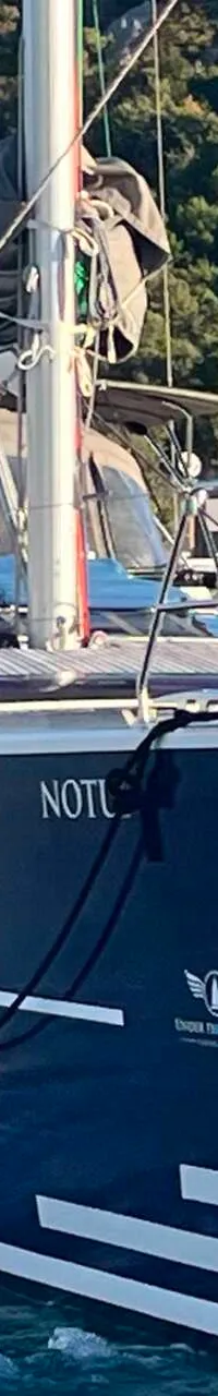 Een donkerblauwe zeilboot met witte strepen en de naam "NOTU" is te zien, varend in water met een groene, beboste…