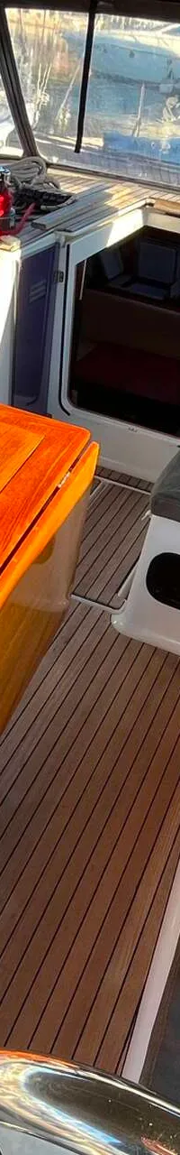 Een houten tafel met een ingebouwde koelkast en bekerhouders staat op een teakhouten dek, omringd door grijze kussens.