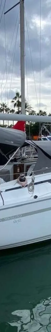 Een witte zeilboot met grijze strepen ligt aangemeerd aan een houten steiger in een haven met palmbomen op de achtergrond.