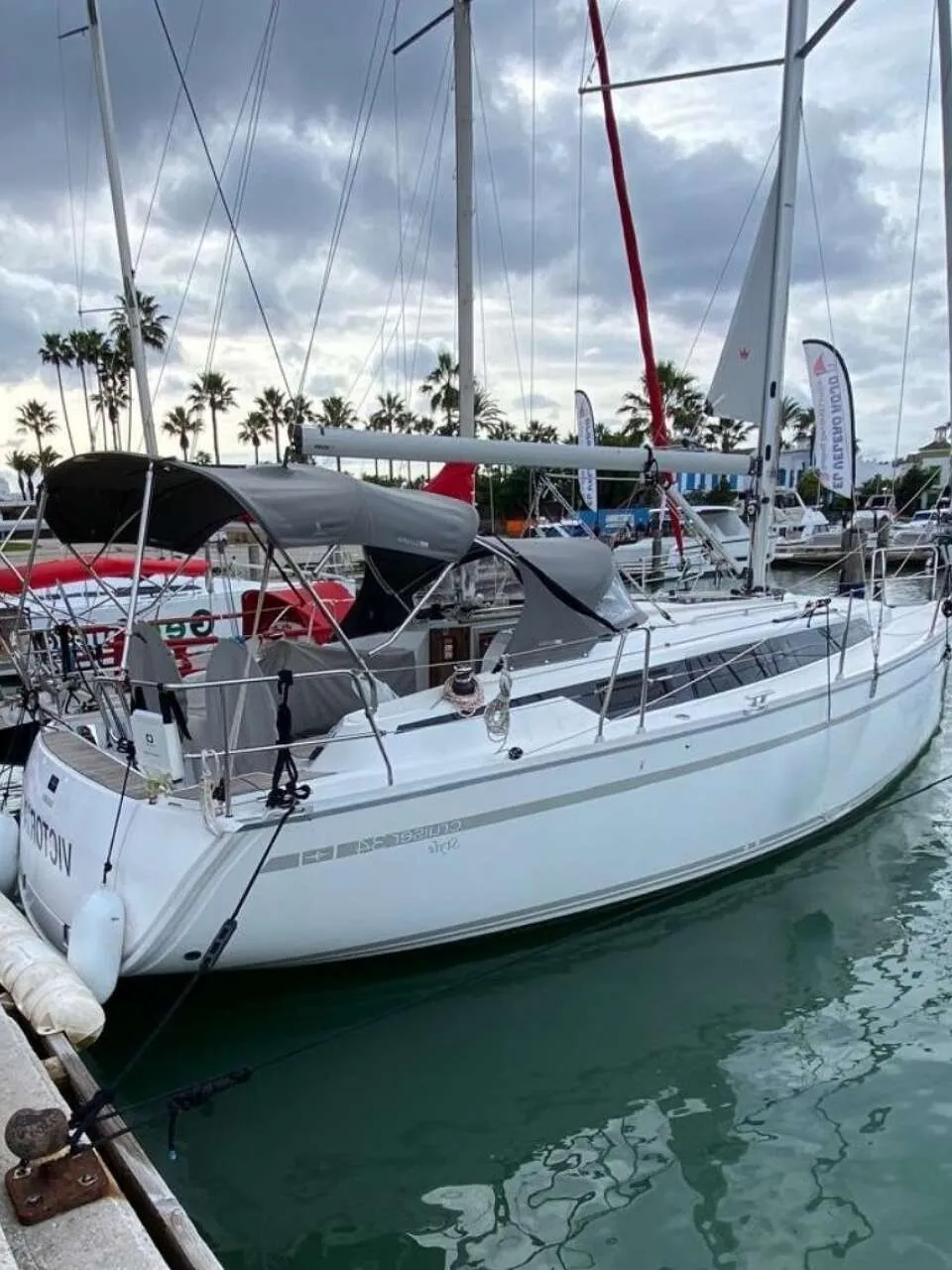 Een witte zeilboot met grijze strepen ligt aangemeerd aan een houten steiger in een haven met palmbomen op de achtergrond.