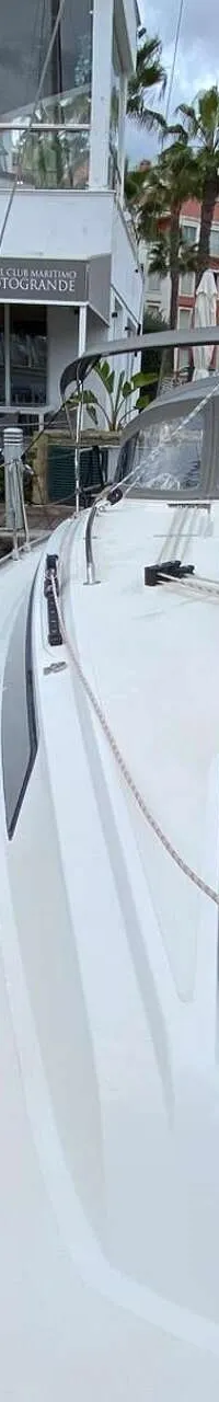 Een witte zeilboot ligt afgemeerd naast een donkerblauwe boot met rode hoezen, in een haven met gebouwen en palmbomen op de…