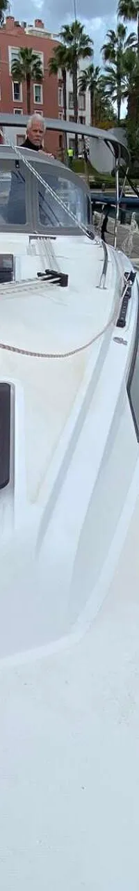 Een witte zeilboot ligt aangemeerd in een haven, met een man in de kuip en gebouwen op de achtergrond. — Bavaria 34 Cruiser