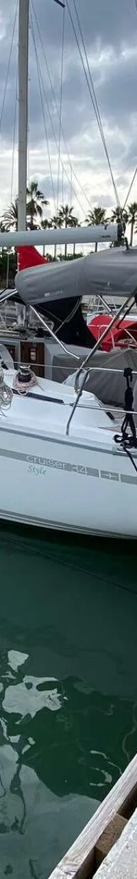 Een witte zeilboot met grijze bimini ligt afgemeerd aan een houten steiger in een haven met palmbomen op de achtergrond.