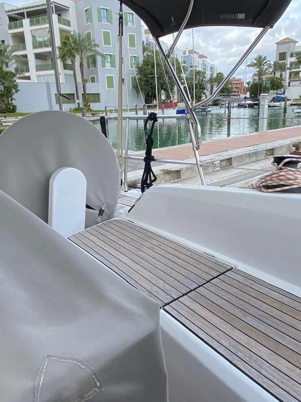 Een boot met een teakdek en afdekhoezen ligt in een jachthaven met gebouwen en palmbomen op de achtergrond.