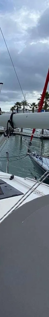 Een bovenaanzicht van een witte zeilboot in een jachthaven met andere boten en palmbomen op de achtergrond.