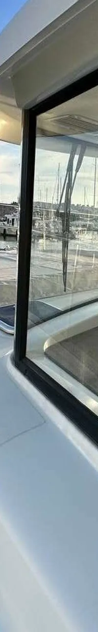 Een witte boot met een reling en stootwillen ligt in een haven met andere boten en bergen op de achtergrond.