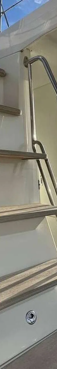 Een trap met houten treden en metalen leuningen leidt naar een bovendek, met uitzicht op water en boten bij zonsondergang.