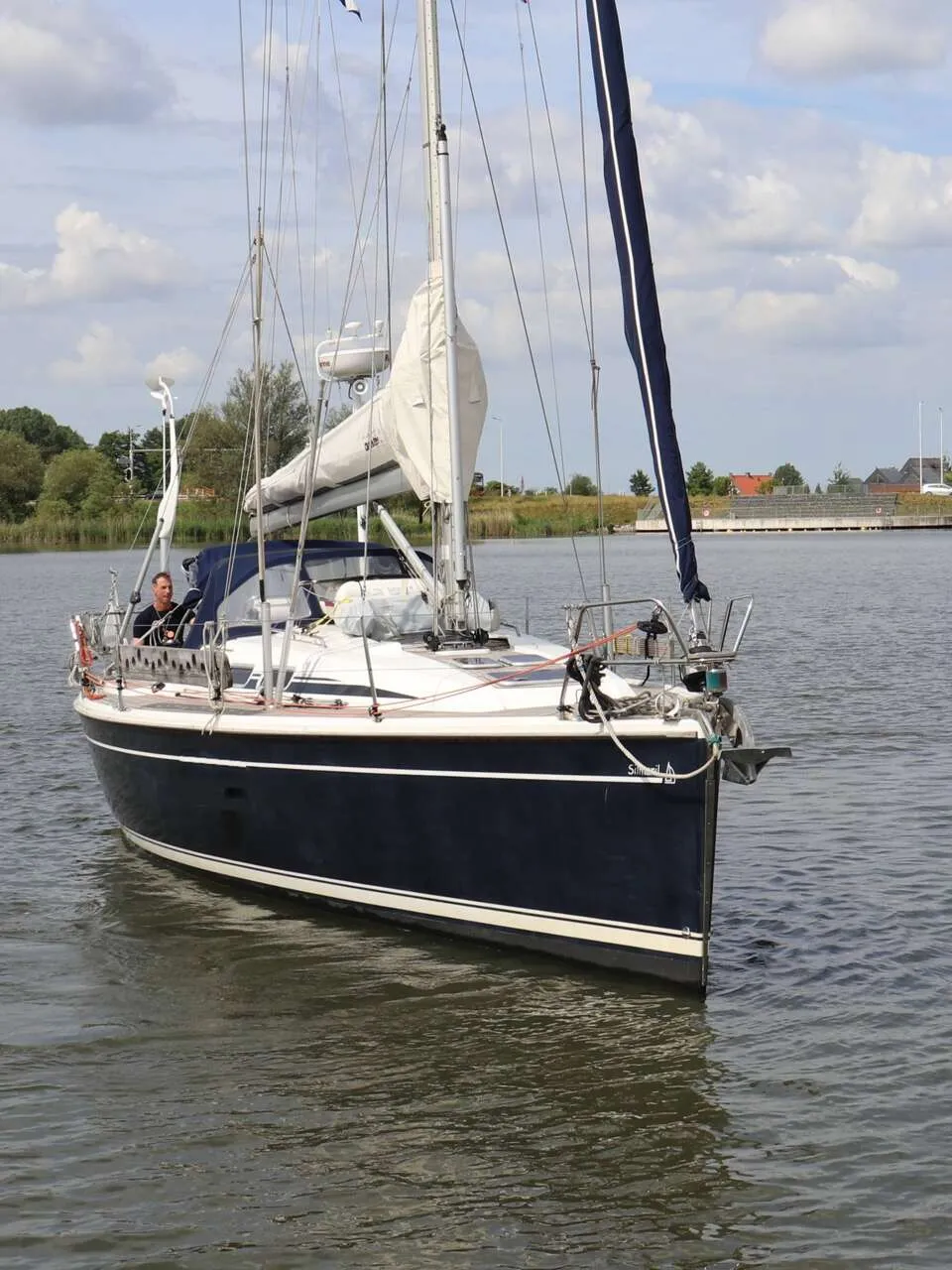 Een donkerblauwe zeilboot met witte strepen vaart op een waterweg met een man aan het roer. — Dehler 39 SQ