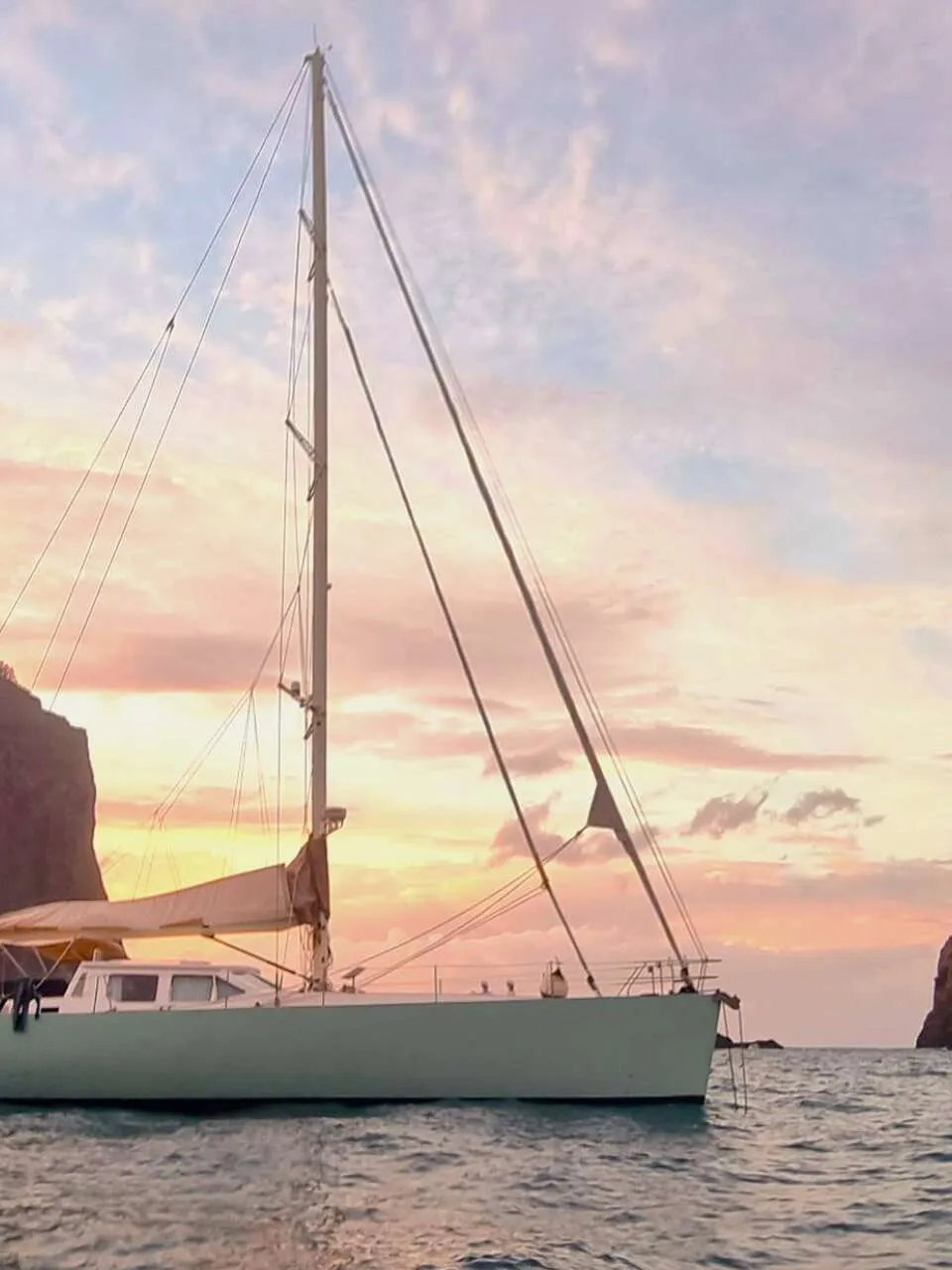 Een lichtgroene zeilboot met de naam "mahi mahi" op de romp vaart op water bij zonsondergang, met rotsachtige kusten op de…