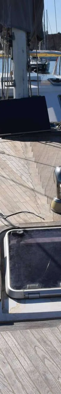 Een houten dek van een zeilboot met een mast, dekramen en reling is zichtbaar, met water en land op de achtergrond. — CRN 63