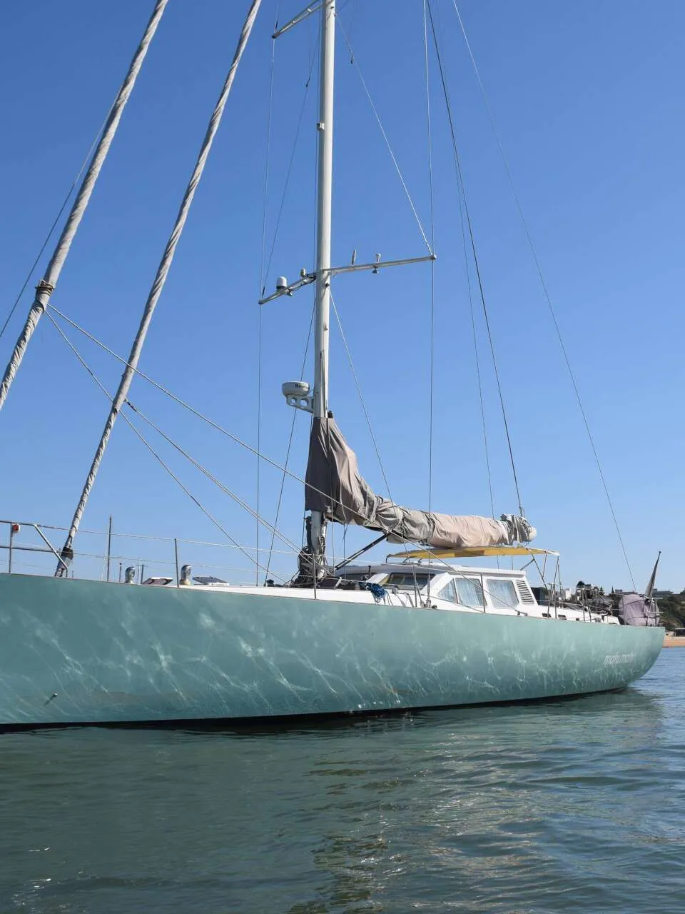 Een lichtgroene zeilboot met opgerolde zeilen en een ankerketting hangt in helder water onder een blauwe lucht, met andere…