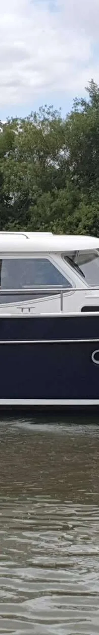 Een donkerblauwe motorboot met witte opbouw en de naam "Grethe" op de spiegel, vaart op water met bomen op de achtergrond.