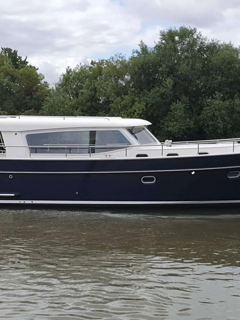 Een donkerblauwe motorboot met witte opbouw en de naam "Grethe" op de spiegel, vaart op water met bomen op de achtergrond.