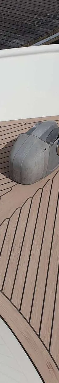 De foto toont het voordek van een boot met een teakdek, een ankerlier en twee bolders met een touw.