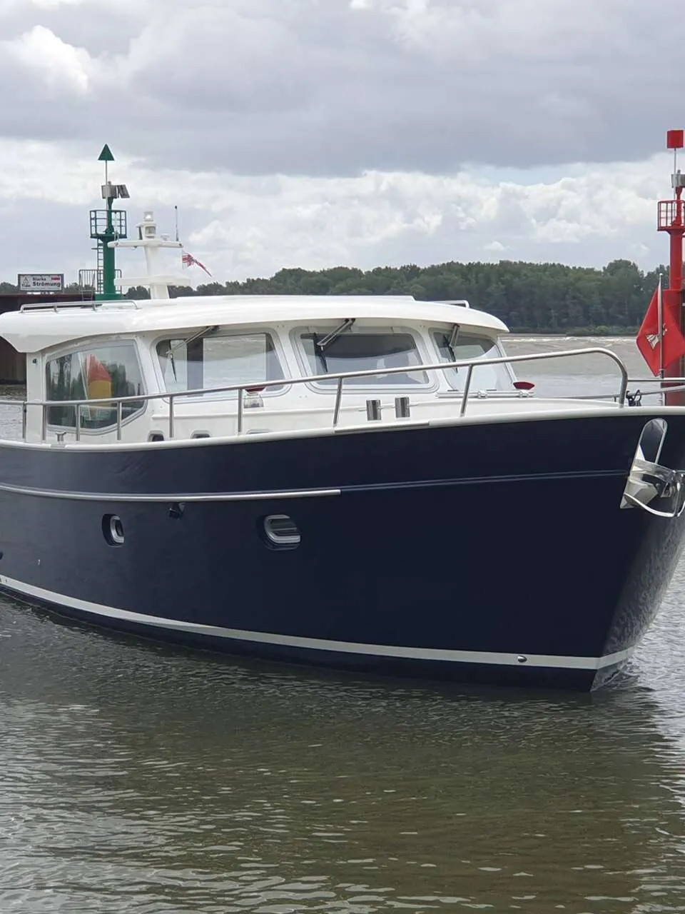 Een donkerblauwe motorboot met witte opbouw en reling vaart op het water, met een rode en groene boei op de achtergrond.