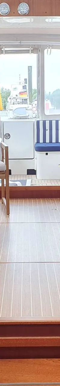Een interieur van een boot met houten vloer en wanden, met een tafel en zitbanken, en een Duitse vlag zichtbaar door de…