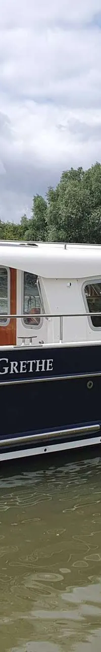 Een blauw-witte motorboot met de naam "GRETHE" vaart op een rivier met een Duitse vlag aan de achtersteven.