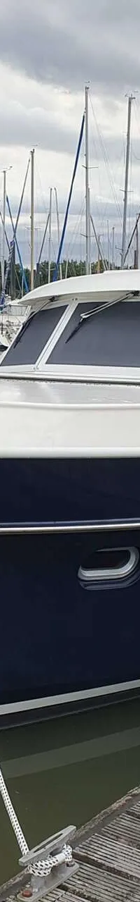 Een blauw-witte motorboot ligt afgemeerd aan een houten steiger, met zeilboten op de achtergrond onder een bewolkte lucht.