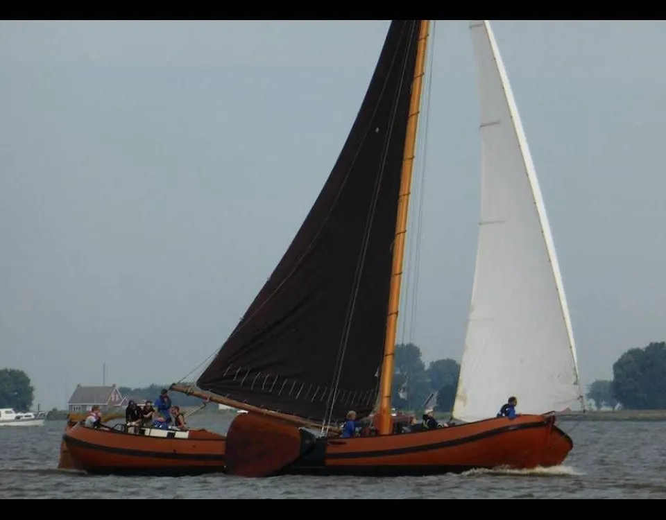Een bruine zeilboot met een zwart grootzeil en een wit voorzeil vaart op het water met mensen aan boord. — Skutsje 19.00