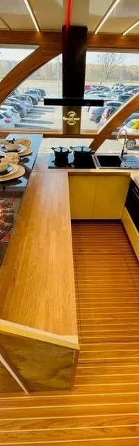 Een interieur van een boot met een keukenblok, zithoek en een groot raam met uitzicht op een parkeerplaats.