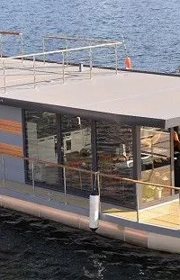 Een moderne woonboot met grote ramen, een plat dak met reling en een houten voordek met reling, ligt in het water.