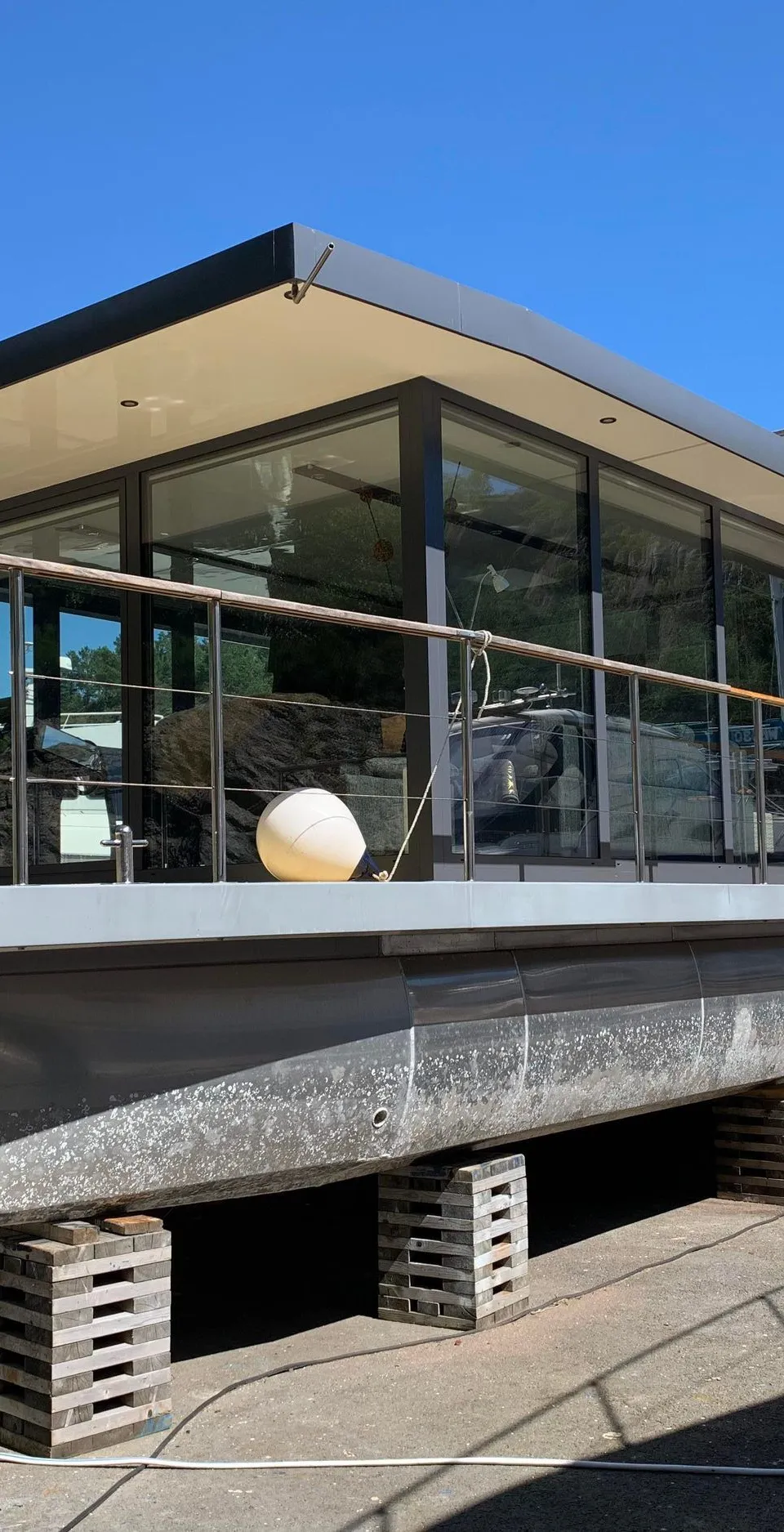 Een moderne woonboot met grote ramen en een plat dak staat op houten blokken op de wal.