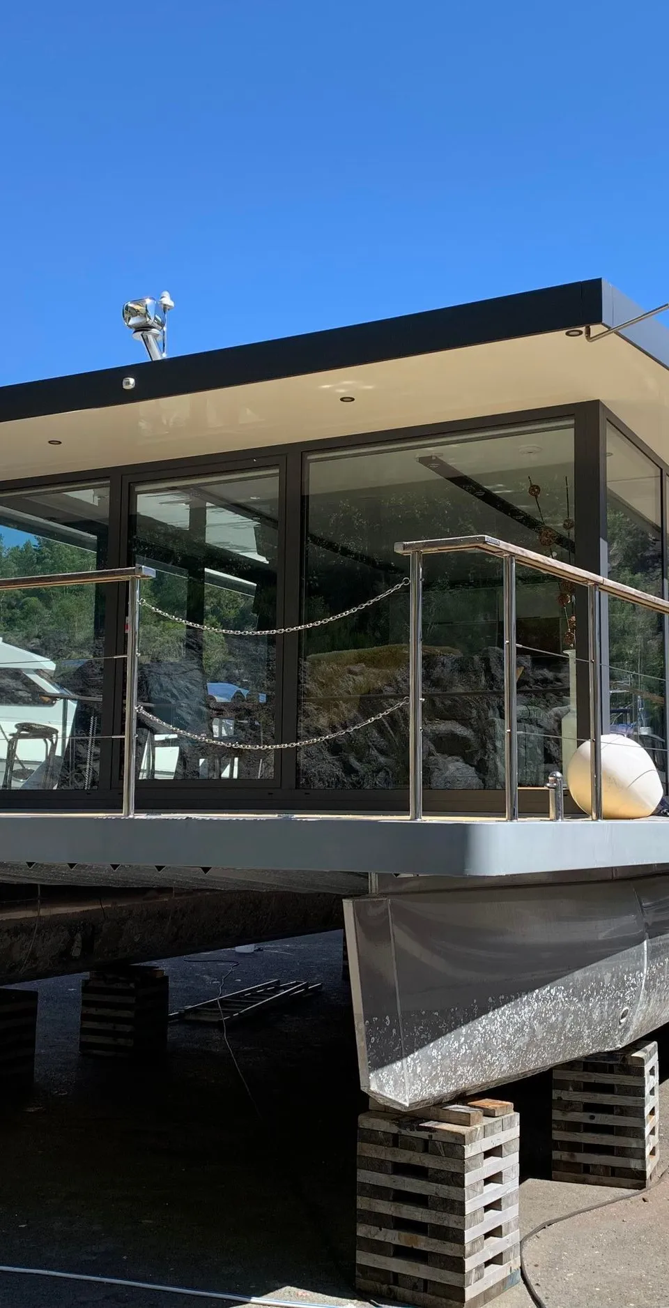 Een moderne woonboot of catamaran met grote ramen en een plat dak staat op houten blokken op het droge.