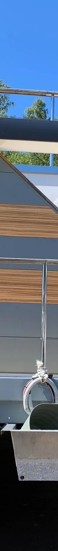 Een moderne woonboot met een grijs-wit-houten exterieur en een dakterras staat op de wal, ondersteund door houten blokken.