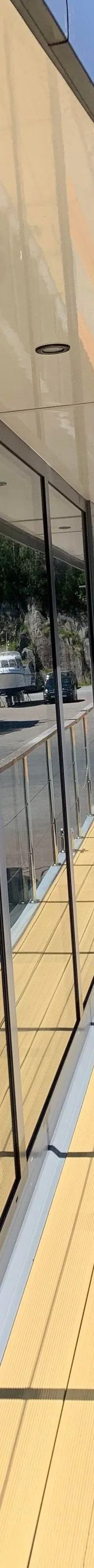 Een gebouw met grote ramen weerspiegelt boten en een rotsachtige heuvel, met een houten dek en boten op de wal.