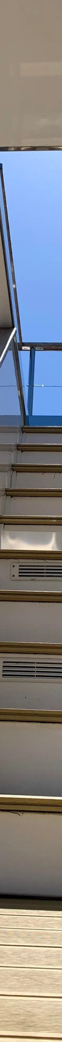 Een trap met witte treden en een glazen wand leidt omhoog, met een blauwe lucht en een deel van een andere boot zichtbaar.