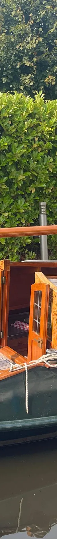Een houten zeilboot met de naam "SAI RAM" ligt aangemeerd naast een groene heg. — Gipon Grundel 800