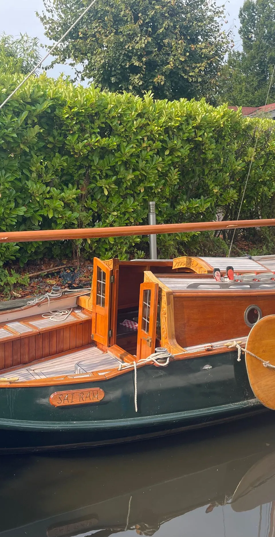 Een houten zeilboot met de naam "SAI RAM" ligt aangemeerd naast een groene heg. — Gipon Grundel 800