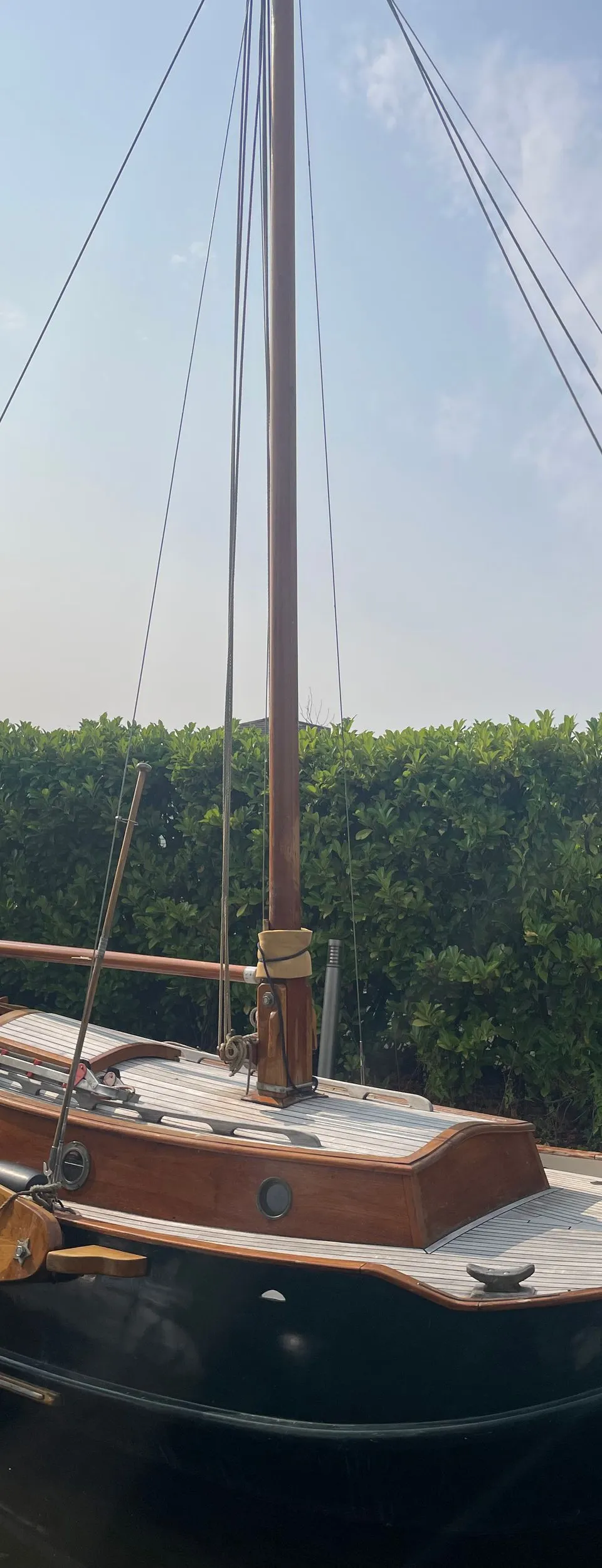 Een donkergroene zeilboot met houten dek en opbouw ligt in het water, met een hek en struiken op de achtergrond.