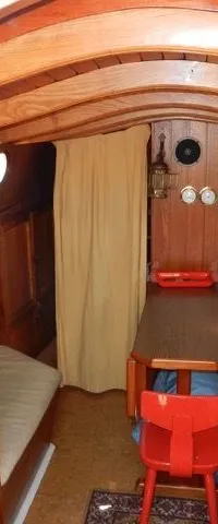 De foto toont het interieur van een boot met houten wanden en plafond, een gootsteen, een tafel met een rode stoel en twee…