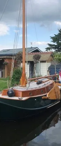 Een donkergroene zeilboot met houten dek en mast ligt in het water, naast een houten vlonder en een rubberboot.
