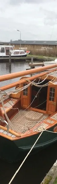 Een donkergroene boot met houten dek en opbouw ligt afgemeerd aan een kade, met andere boten op de achtergrond.
