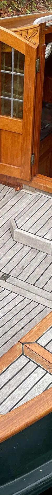 Een houten zeilboot met een donkergroene romp ligt aangemeerd, met de kajuitdeuren open en een zichtbare kombuis.