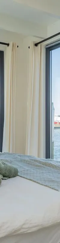 Een bed met witte lakens, kussens en opgerolde handdoeken, met uitzicht op een jachthaven met boten.