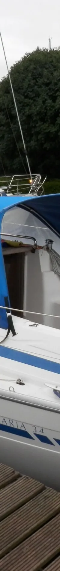 Een witte zeilboot met blauwe strepen en blauwe kappen ligt aangemeerd aan een houten steiger, met "BAVARIA 34" op de romp.