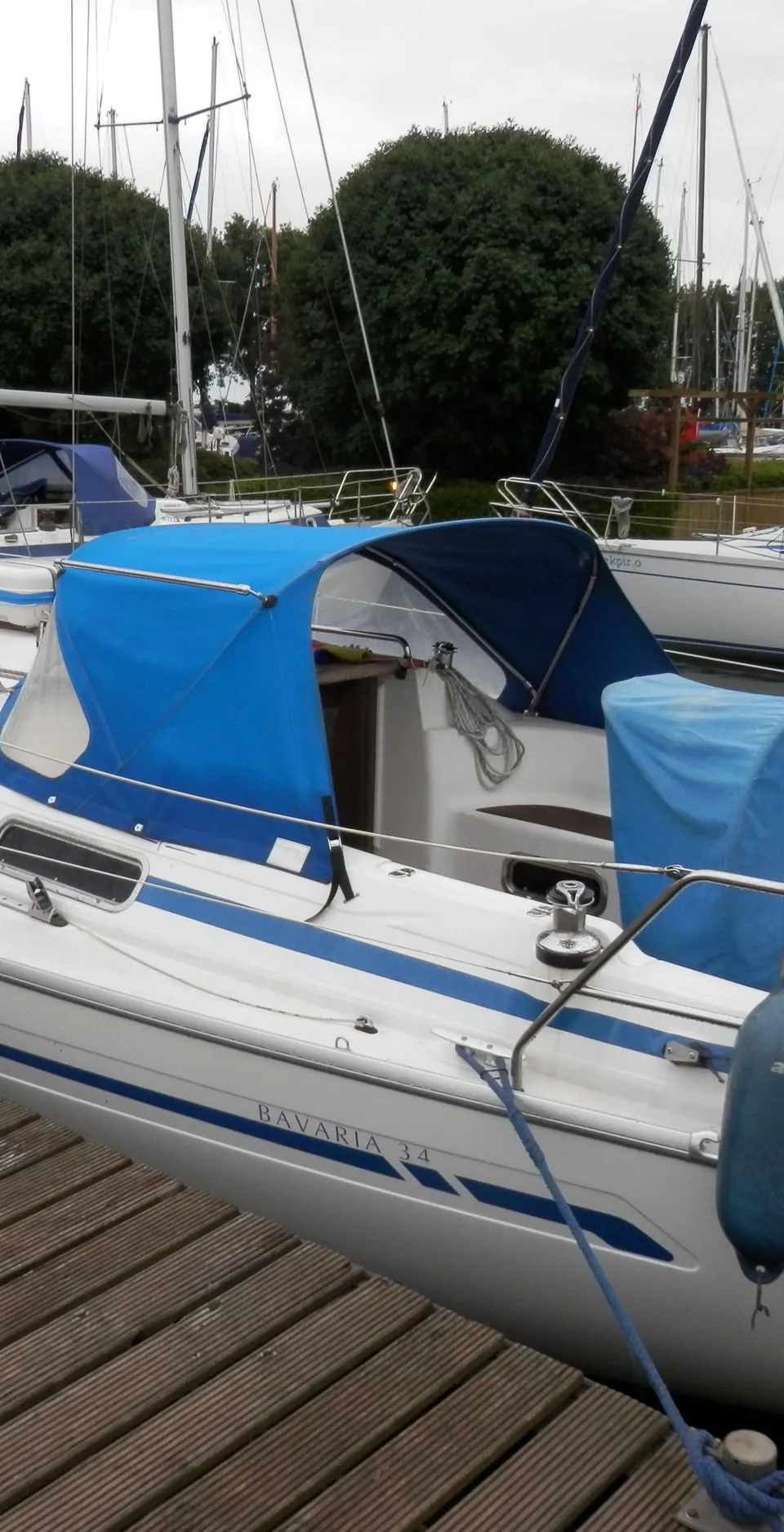 Een witte zeilboot met blauwe strepen en blauwe kappen ligt aangemeerd aan een houten steiger, met "BAVARIA 34" op de romp.