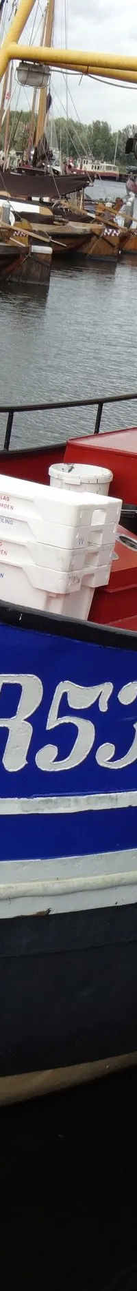Een blauw-rood schip met de aanduiding "WR53" ligt afgemeerd aan een kade, met andere boten en gebouwen op de achtergrond.