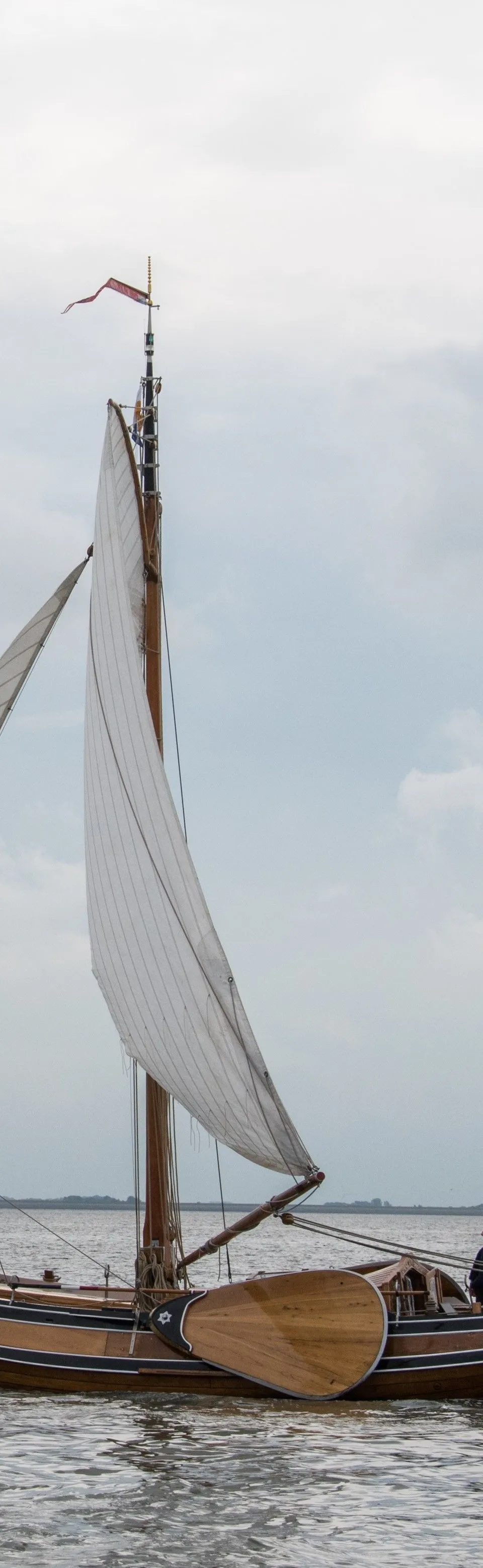 Een houten zeilboot met twee zeilen en twee personen aan boord vaart op water onder een bewolkte hemel. — Boeier Lantinga 900